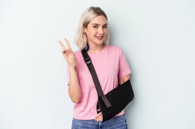 Kırık elli beyaz bir kadın mavi arka planda izole edilmiş neşeli ve kaygısız parmaklarıyla barış sembolü gösteriyor..