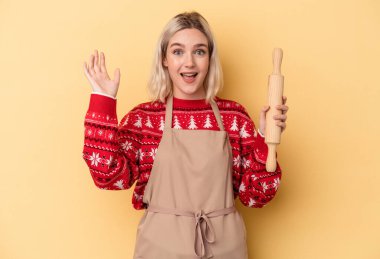 Beyaz tenli genç fırıncı kadın, sarı arka planda izole bir şekilde Noel için kurabiye yapıyor. Hoş bir sürpriz alıyor, heyecanlı ve el kaldırıyor..