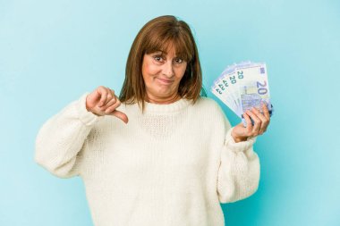 Middle age caucasian woman holding bank notes isolated on blue background showing a dislike gesture, thumbs down. Disagreement concept.