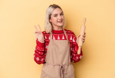 Kafkasyalı genç fırıncı kadın Noel için kurabiye yapıyor sarı arka planda izole edilmiş neşeli ve tasasız parmaklarıyla barış sembolü gösteriyor..