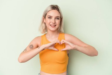 Yeşil arka planda izole edilmiş genç beyaz kadın gülümsüyor ve elleriyle kalp şeklini gösteriyor..