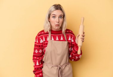 Beyaz tenli fırıncı kadın Noel için kurabiye yapıyor. Sarı arka planda izole edilmiş. Omuzlarını silkiyor ve gözleri açık..
