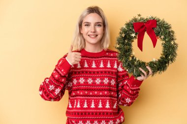 Sarı arka planda izole edilmiş bir Noel çelengi tutan genç beyaz kadın gülümsüyor ve başparmağını kaldırıyor.
