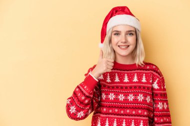 Genç beyaz kadın Noel 'i kutluyor sarı arka planda izole edilmiş gülümsüyor ve başparmağını kaldırıyor.