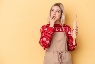 Beyaz tenli fırıncı kadın Noel için kurabiye yapıyor. Sarı arka planda izole edilmiş. Bir şeyin fotokopi odasına baktığını düşünüyor..