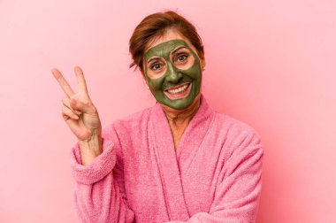 Middle age caucasian woman wearing a facial mask isolated on pink background joyful and carefree showing a peace symbol with fingers.