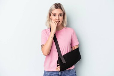 Kırık elli beyaz bir kadın mavi arka planda izole edilmiş tırnak yiyor, gergin ve endişeli..