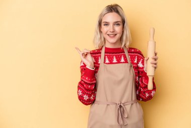 Beyaz tenli genç fırıncı kadın Noel için kurabiye yapıyor sarı arka planda izole edilmiş bir şekilde gülümsüyor ve boş alanda bir şeyler gösteriyor..