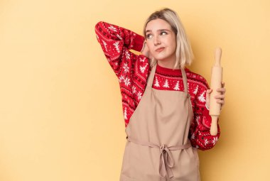 Kafkasyalı genç fırıncı kadın Noel için kurabiye yapıyor sarı arka planda izole edilmiş kafasının arkasına dokunuyor, düşünüyor ve bir seçim yapıyor..