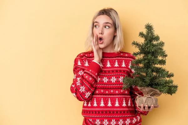 年轻的高加索女人抱着一棵被黄色背景隔离的小圣诞树 正在说着一个秘密的 火爆的刹车消息 并将它抛在一边 — 图库照片