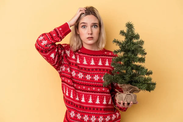 若い白人女性が黄色の背景に孤立した小さなクリスマスツリーを持っているショックを受けて 彼女は重要な会議を覚えています — ストック写真