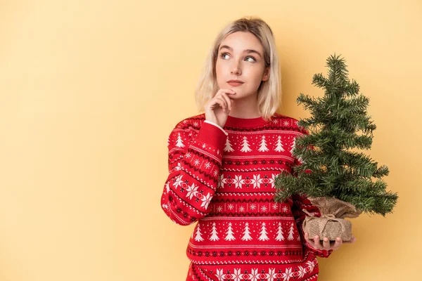 年轻的高加索女人抱着一棵被黄色背景隔开的小圣诞树 侧视着 脸上带着怀疑和怀疑的表情 — 图库照片