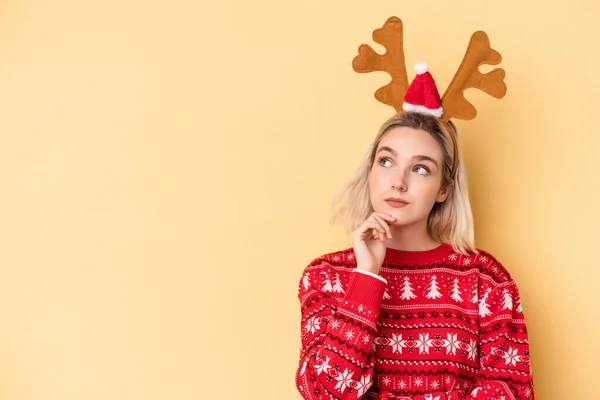 年轻的高加索女人头戴一顶黄底的圣诞驯鹿帽 面带怀疑和怀疑的表情侧视着四周 — 图库照片