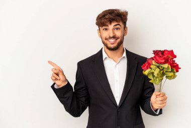 Genç melez adam elinde gül buketi tutarken beyaz arka planda izole edilmiş gülümseyen ve işaret eden boş alanda bir şey gösteriyor..