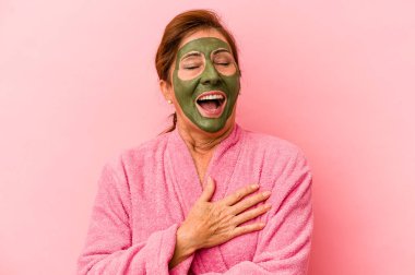 Middle age caucasian woman wearing a facial mask isolated on pink background laughs out loudly keeping hand on chest.