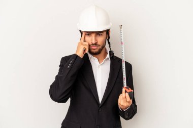 Miğfer takan ve taksimetre tutan genç bir mimar gri arka planda izole edilmiş, parmağıyla tapınağı işaret ediyor, düşünüyor ve bir göreve odaklanıyor..