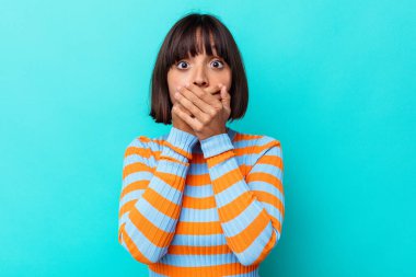 Mavi arka planda izole edilmiş genç melez kadın elleri endişeli bir şekilde ağzı örtüyor..