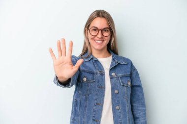 Mavi arka planda izole edilmiş genç beyaz kadın gülümseyerek beş numarayı parmaklarıyla gösteriyor..