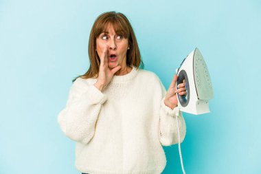 Middle age caucasian woman holding a iron isolated on blue background is saying a secret hot braking news and looking aside