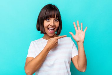 Mavi arka planda izole edilmiş genç melez kadın gülümseyerek beş numarayı parmaklarıyla gösteriyor..