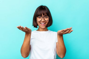 Mavi arka planda soyutlanmış genç melez kadın kollarıyla ölçeği yapıyor, mutlu ve kendinden emin hissediyor..