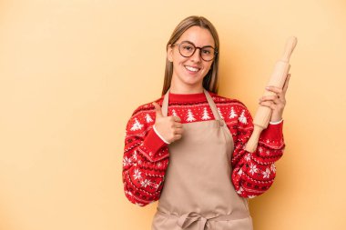Genç fırıncı kadın Noel için kurabiye yapıyor sarı arka planda izole edilmiş gülümsüyor ve başparmağını kaldırıyor.