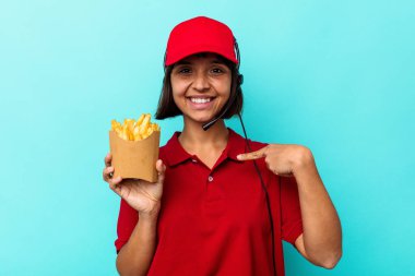 Genç melez ırk fast food restoranı çalışanı elinde patates kızartması, mavi arka planda izole edilmiş, gömlek fotokopi alanını işaret eden, gururlu ve kendinden emin.