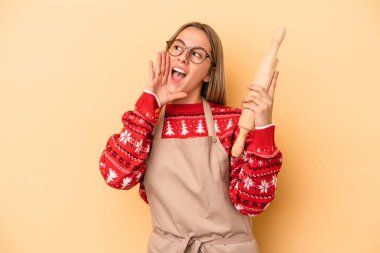 Genç fırıncı kadın Noel için kurabiye yapıyor sarı arka planda izole edilmiş bağırıyor ve avuç içini açık tutuyor..