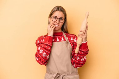 Genç fırıncı kadın Noel için kurabiye yapıyor. Sarı arka planda izole edilmiş tırnak yiyor, gergin ve endişeli..