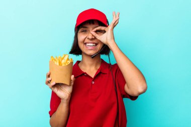 Genç melez kadın fast food restoranı çalışanı elinde kızartmalarla mavi arka planda izole edilmiş heyecanlı jestler yapıyor..