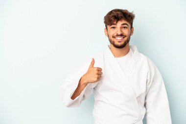 Mavi arka planda karate yapan genç melez adam gülümsüyor ve başparmağını kaldırıyor.