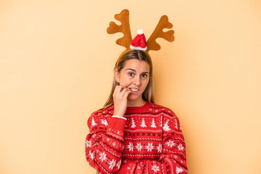 Sarı arka planda izole edilmiş, gergin ve endişeli bir şekilde tırnak yiyen, Noel geyiği şapkası takan beyaz bir kadın..