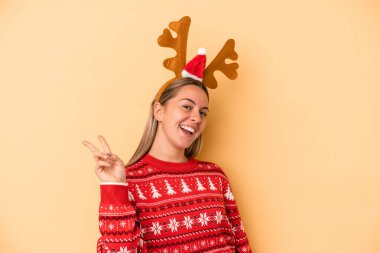 Sarı arka planda izole edilmiş, Noel geyiği şapkası takan beyaz, genç bir kadın. Neşeli ve tasasız, parmaklarıyla barış sembolü gösteriyor..