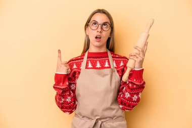 Genç fırıncı kadın Noel için kurabiye yapıyor. Sarı arka planda izole edilmiş, ağzı açık, üst tarafı işaret ediyor..