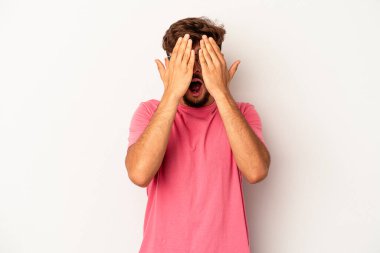 Genç melez adam gri arka planda izole edilmiş. Yüzünün yarısını avuç içiyle kaplayarak eğleniyor..