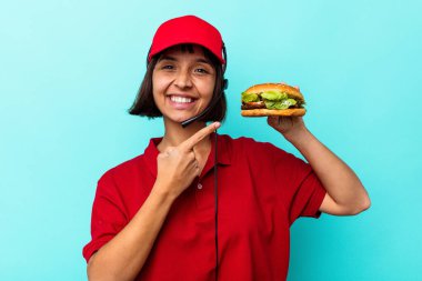 Genç melez kadın fast food restoranı çalışanı mavi arka planda izole edilmiş bir burger tutuyor.