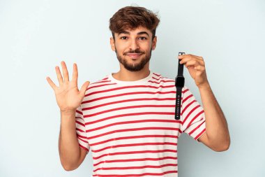 Mavi arka planda izole edilmiş bir kol saati tutan genç melez adam gülümseyerek beş numarayı parmaklarıyla gösteriyor..