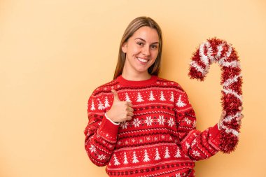 Sarı arka planda izole edilmiş büyük bir Noel sopası tutan genç beyaz kadın gülümsüyor ve başparmağını kaldırıyor.