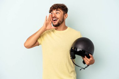 Mavi arka planda izole edilmiş motosiklet kaskı tutan melez genç adam bağırıyor ve avuç içini açık tutuyor..