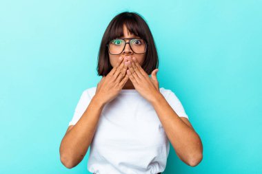 Mavi arka planda izole edilmiş genç melez kadın elleri endişeli bir şekilde ağzı örtüyor..