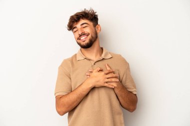 Genç melez adam gri arka planda soyutlanmış gülüyor, ellerini kalbe koyuyor, mutluluk kavramını koruyor..