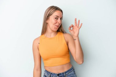 Kafkasyalı genç bir kadın mavi arka planda izole edilmiş bir şekilde bir göz kırpıyor ve elleriyle iyi bir jest yapıyor..