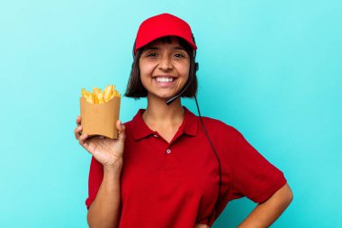 Genç melez kadın fast food restoranı çalışanı elinde kızartmalarla mavi arka planda mutlu, güler yüzlü ve neşeli..