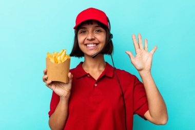 Genç melez ırk fast food restoranı çalışanı elinde kızartmalarla mavi arka planda izole bir şekilde gülümseyerek beş numarayı parmaklarıyla gösteriyor..