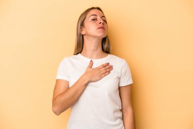 Sarı arka planda izole edilmiş genç beyaz kadın yemin ediyor, elini göğsüne koyuyor..