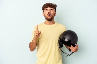 Mavi arka planda izole edilmiş motosiklet kaskı tutan genç melez adam ağzı açık bir şekilde üst tarafı işaret ediyor..