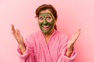Middle age caucasian woman wearing a facial mask isolated on pink background receiving a pleasant surprise, excited and raising hands.