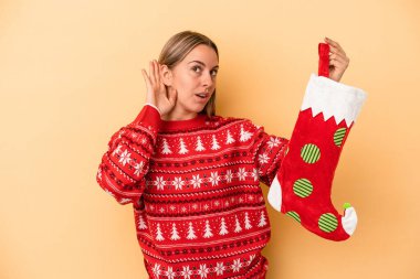Sarı arka planda izole edilmiş bir elf çorabı tutan genç beyaz kadın bir dedikoduyu dinlemeye çalışıyor..