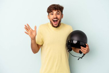 Mavi arka planda izole edilmiş motosiklet kaskı tutan genç bir ırk hoş bir sürpriz alıyor, heyecanlı ve el kaldırıyor..