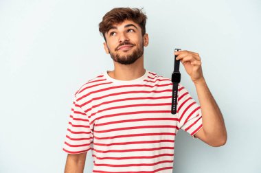 Genç melez bir adam mavi arka planda izole edilmiş bir kol saati tutuyor. Amaçlarına ve amaçlarına ulaşmayı hayal ediyor.